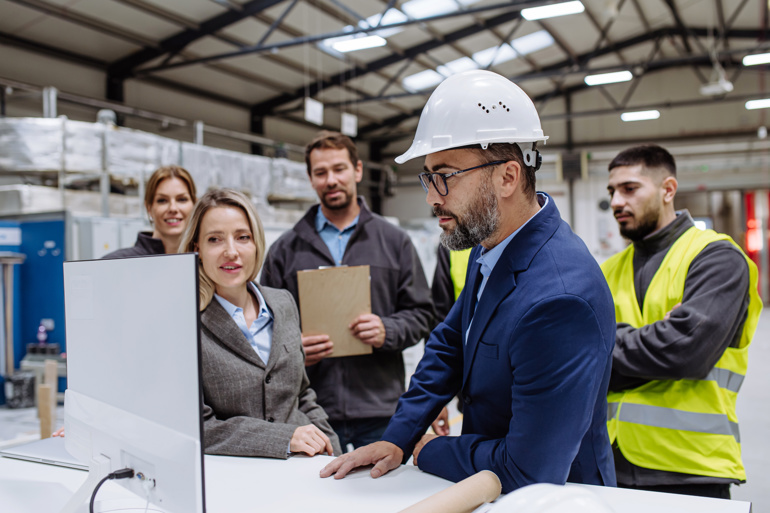 A project management professional in a construction site hard hat showing a team of project professionals some work on a computer screen